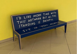 Shannon Finnegan, Blue Bench: I’d Like More Time With This Artwork But All This Standing Is Painful. Sit If You Agree, 2021 [image description: a dark blue bench against a yellow wall. Writing in capitalized, white letters reads “I’d like more time with this artwork but all this standing Is painful.” on the back. The words “sit if you agree” are on the seat.]